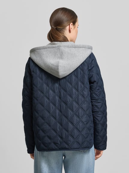 Kobiece modelka nosi Regular Fit kurtka z efektem pikowania z kapturem model 'Fenya' od Soyaconcept, wpuszczane kieszenie, listwa guzikowa, kolor granatowy, długość tyłu 65 cm, szerokość ramion 40 cm, szerokość w rozmiarze S 54 cm, długość rękawów 65 cm, widok całej sylwetki z tyłu.