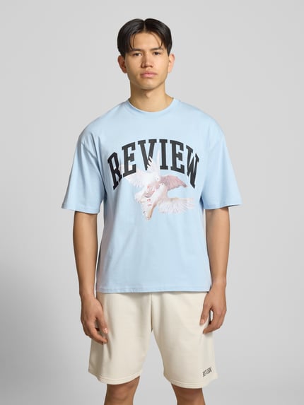 Männermodel trägt T-Shirt mit Logo- und Motiv-Print von REVIEW in Hellblau, Reine Baumwolle, Loose-Fit, gerippter Rundhalsausschnitt, Ganzkörperansicht von vorne. Rückenlänge bei Größe M: 70 cm.