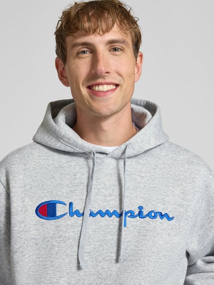 Männermodel trägt grauen Hoodie mit Label-Stitching von Champion aus Baumwoll-Mix, lockerer Schnitt, Nahaufnahme Oberkörper.