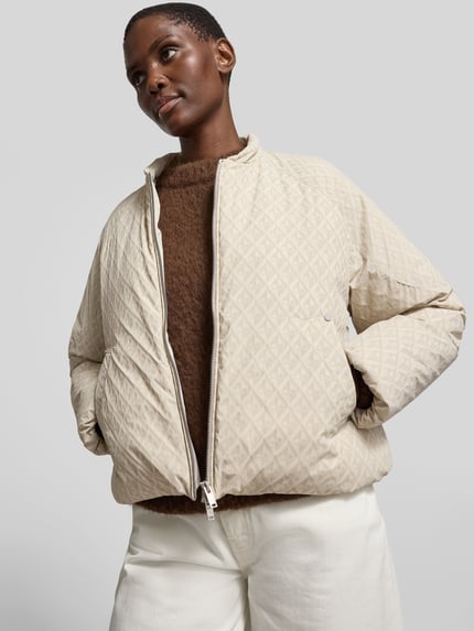 Kobiece model prezentuje Regular Fit Jacke mit Allover-Logo-Print od Moose Knuckles, jasny beż, wzór geometryczny, krój regular fit, elastyczna mieszanka materiałów, kaptur ze ściągaczem, dwukierunkowy zamek, długość tyłu w rozmiarze S: 60 cm, szerokość w rozmiarze S: 59 cm, długość rękawa w rozmiarze S: 63 cm, ujęcie całej sylwetki z przodu.