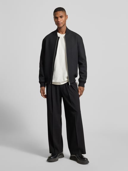 Männermodel trägt Wide Fit Hose mit Bügelfalten Modell 'Leek' von Drykorn, schwarzer Material-Mix mit Viskose-Anteil, Ganzkörperansicht von vorne.