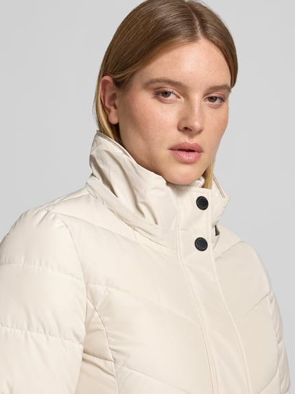 Frauenmodel trägt Steppmantel mit Kapuze von NAUTGÅRD 'NEBULA' in creme, Struktur gesteppt, Nahaufnahme Oberkörper, Regular Fit, verdeckter Reißverschluss, Reißverschlusstaschen, Kontext: Rückenlänge 108 cm, Schulterbreite 42 cm, Brustweite 37 cm, Ärmellänge 67 cm.