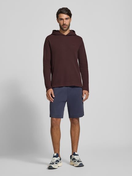 Männermodel trägt Relaxed Fit Sweatshirt aus Baumwoll-Mix Modell 'Waffle LS' von BOSS in Dunkelrot, kombiniert mit blauen Shorts, Ganzkörperansicht von vorne, Herren Hoodie von BOSS.