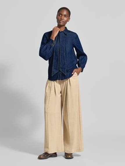 Modelka nosi Regular Fit Hemdbluse aus reiner Baumwolle in Denim-Optik von Emily Van den Bergh, tkanina stylizowana na denim, krój regular fit, kołnierzyk koszulowy, ozdobna kokarda, długi rękaw, bawełna, widoczne kontrastowe szwy, długość tyłu 64 cm, długość rękawów 59 cm, Ganzkörperansicht von vorne.