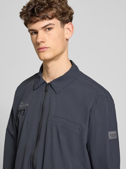 Männermodel trägt Regular Fit Hemdjacke mit Logo Modell 'HALO MOVE' von HALO, wykładany kołnierz, zamek błyskawiczny, długość tyłu 66 cm, długość rękawów 61 cm, kolor ciemnoszary, struktura gładka, widoczne logo, Nahaufnahme górnej części ciała.