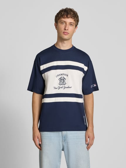 Männermodel trägt Relaxed Fit T-Shirt aus reiner Baumwolle von Champion, blau-weiß mit Logo-Print und Motiv-Stitching, Ganzkörperansicht von vorne. Rückenlänge bei Größe M 70 cm, Rundhalsausschnitt.