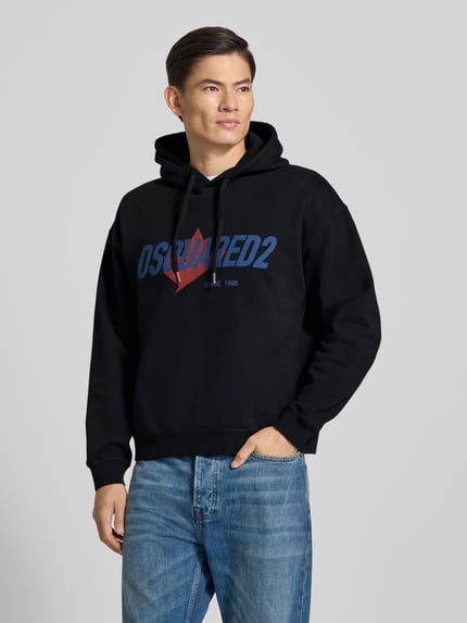 Männermodel trägt Relaxed Fit Hoodie aus reiner Baumwolle von Dsquared2, schwarz mit blau-rotem Label-Detail, Ganzkörperansicht von vorne.