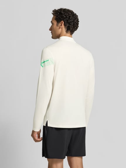 Męski model nosi bluzkę z długim rękawem Model 'ICON' od ASICS TIGER, prosty krój, nadruk z logo, kołnierz zapinany, czarna i biała kolorystyka, długość tyłu w rozmiarze M: 72 cm, widok całej sylwetki od tyłu.