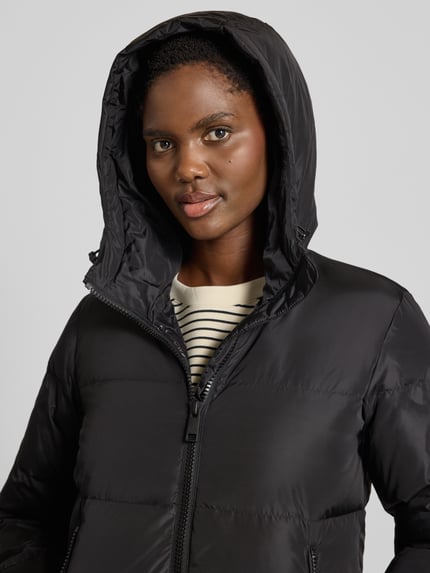 Frauenmodel trägt Steppmantel mit Zweiwege-Reißverschluss von Armani Exchange, schwarze Farbe, glatte Oberfläche, Nahaufnahme Oberkörper. Gerade Passform, Kapuze mit Tunnelzug, Reißverschlusstaschen, Storm Cuffs, Rückenlänge bei Größe S 100 cm, Schulterbreite 43 cm, Brustweite 55 cm, Ärmellänge 63 cm.