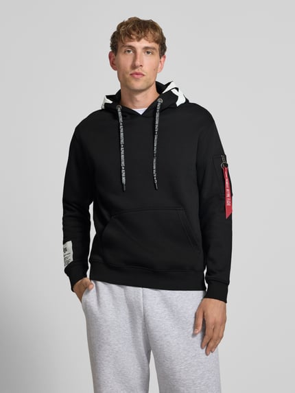 Männermodel trägt Regular Fit Hoodie aus Baumwoll-Mix mit Label-Patch von Alpha Industries in Schwarz, Känguru-Tasche, Ganzkörperansicht von vorne, Rückenlänge 67 cm, Ärmellänge 63 cm, Ärmel-Tasche, Kapuze mit Tunnelzug.