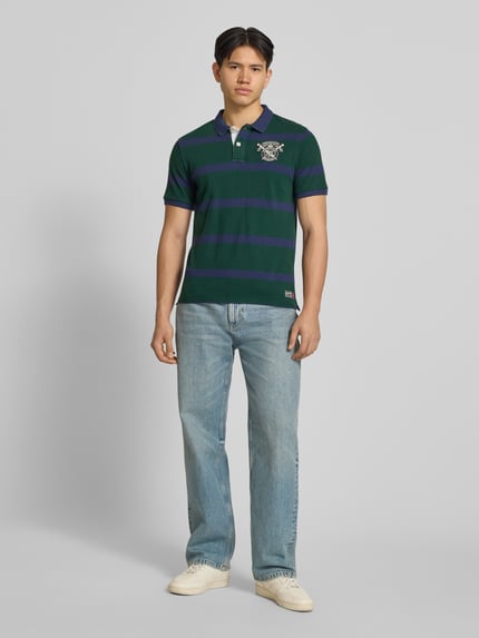 Männermodel trägt Slim Fit Poloshirt mit Label-Stitching und Knopfleiste von Superdry, Reine Baumwolle, Rückenlänge 70 cm, Ganzkörperansicht von vorne.