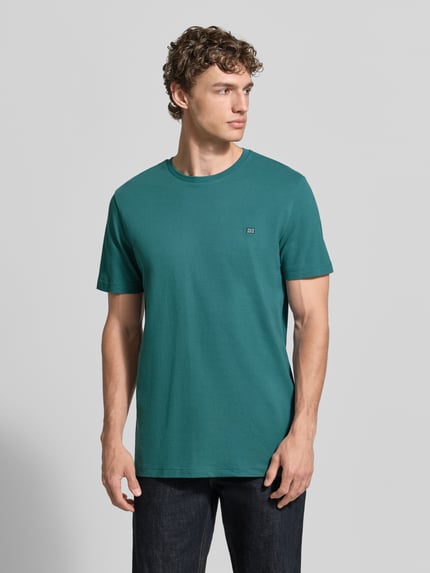Męski model ma na sobie T-Shirt aus Baumwolle mit Rundhalsausschnitt od Christian Berg Men w kolorze zielonym, krój regular fit, detal z logo przy dekolcie, czysta bawełna, długość tyłu w rozmiarze M: 71 cm, widok całej sylwetki z przodu.