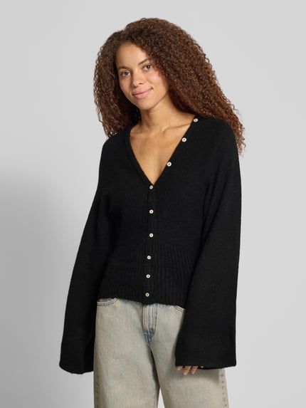 Vrouwenmodel draagt Regular Fit gebreid jack met wol van Gina Tricot, zwarte elastische materiaalmix, ribboorden, knoopsluiting, Met lange mouwen, ruglengte maat S 53 cm, mouwlengte maat S 49 cm, volledig design, voorkantaanzicht.