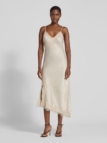 Frauenmodel trägt Maxikleid mit Spitzenbesatz von VERO MODA in glänzendem Beige, Satinstoff, Spitzenmuster, asymmetrischer Saum, Ganzkörperansicht von vorne.