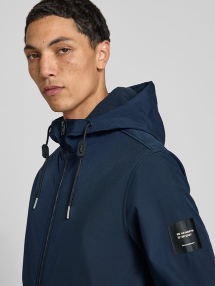 Männermodel trägt Regular Fit Jacke mit Logo-Detail von JACK & JONES in Dunkelblau, glatte Textur, Nahaufnahme Oberkörper. Softshell, Kapuze mit Tunnelzug, Eingrifftaschen mit Reißverschluss, elastische Abschlüsse, Rückenlänge 66 cm, Schulterbreite 46 cm, Brustweite 53 cm, Ärmellänge 67 cm.