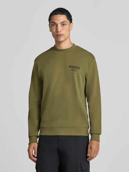 Męski model nosi Regular Fit Sweatshirt aus reiner Baumwolle model 'Nespon' od HUGO, oliwkowa z czarnym nadrukiem, bluza bawełniana, widok całej sylwetki z przodu, kolekcja HUGO Blue.