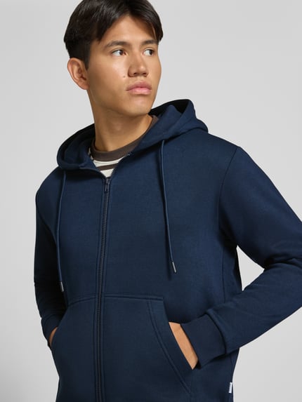 Męski model nosi bluza rozpinana z mieszanki bawełny o kroju regular fit Model 'BRADLEY' od JACK & JONES, kolor granatowy, kieszeń kangurka, kaptur z tunel, elastyczne wykończenia, długość tyłu w rozmiarze M: 67 cm, długość rękawów w rozmiarze M: 68 cm, półzbliżenie przód.