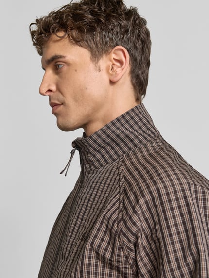Männermodel trägt Bomberjacke mit Raglanärmeln von Forét in Braun mit Karomuster, Stehkragen, sichtbaren Nähten, Nahaufnahme Oberkörper. Rückenlänge 64 cm, Schulterbreite 42 cm, Ärmellänge 62 cm, lockere Passform, Zweiwege-Reißverschluss, elastische Abschlüsse.