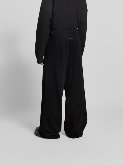 Mannelijk model draagt Wide Fit stoffen broek met achterzak van MM6 Maison Margiela, puur katoen, wide fit, elastische band, open zoom, binnenbeenlengte maat M 82 cm, bandwijdte maat M 38 cm, geheel in zwart, Ganzkörperansicht Rückansicht.