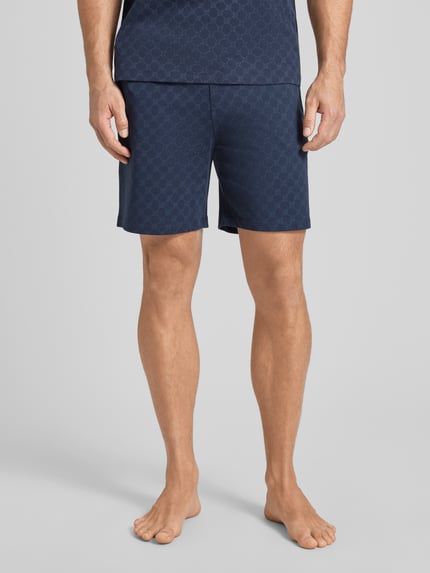Mężczyzna nosi Shorts aus Baumwolle mit Logo-Muster von JOOP! Collection, bawełna, krój regular fit, wzór z logo, elastyczny pas, szerokość w rozmiarze M 42 cm, długość nogawki wewnętrznej w rozmiarze M 18 cm, granatowy, widok z przodu, ujęcie całej sylwetki.