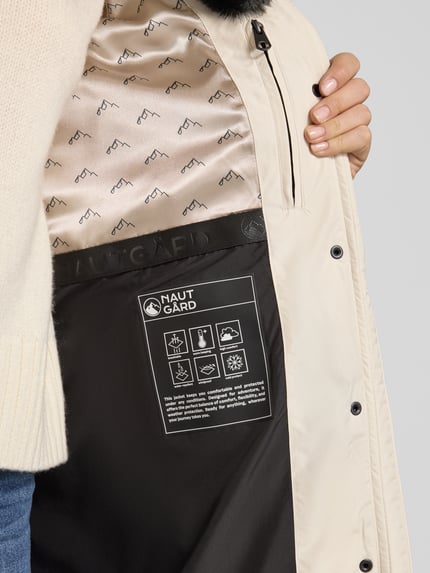 Frauenmodel trägt Steppmantel mit Kapuze von NAUTGÅRD 'NEBULA', beige Außenstoff, Innenfutter mit Markenmuster, Nahaufnahme Oberkörper. Damen Steppmantel Regular Fit, verdeckter Reißverschluss, Reißverschlusstaschen, Rückenlänge 108 cm, Schulterbreite 42 cm, Brustweite 37 cm, Ärmellänge 67 cm.