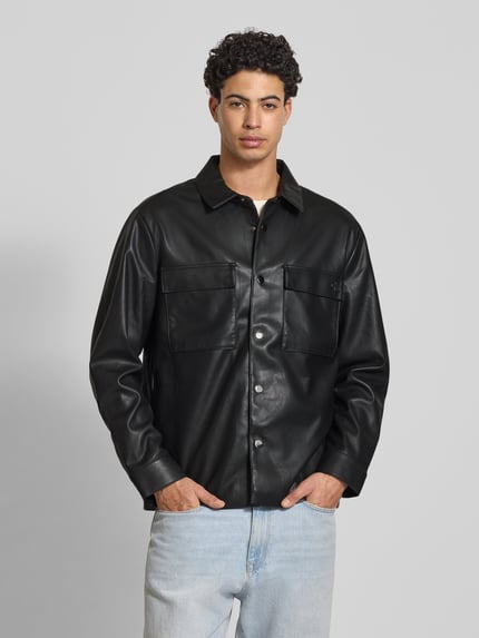 Męski model nosi Regular Fit Hemdjacke in Leder-Optik von Calvin Klein Jeans, czarny materiał skóropodobny, krój regular fit, wykładany kołnierz, kieszenie na piersi, listwa guzikowa, sportowe mankiety, długość tyłu w rozmiarze M 73 cm, szerokość w rozmiarze M 50 cm, długość rękawów w rozmiarze M 65 cm, jasnoniebieskie jeansy, gładka tekstura, widok całej sylwetki z przodu.