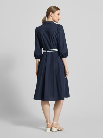 Frauenmodel trägt Hemdblusenkleid mit V-Ausschnitt und Bindegürtel von More & More in Dunkelblau, Baumwoll-Elasthan-Mix, A-Linie, Midikleid, Knopfleiste, Rückenlänge 109 cm, Brustweite 47 cm, Ärmellänge 49 cm, Ganzkörperansicht Rückansicht.