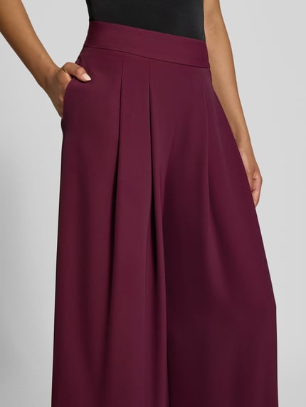 Frauenmodel trägt Wide Leg Stoffhose mit Reißverschlusstaschen von Rinascimento in dunklem Bordeaux, glatte Textur, Nahaufnahme Oberkörper. Material-Mix mit Stretch-Anteil, elastischer Bund, seitliche Reißverschlusstaschen, Bundweite bei Größe S 37 cm, Innenbeinlänge bei Größe S 82 cm.