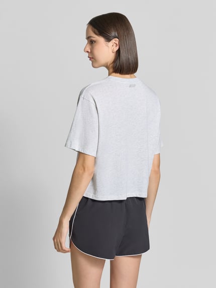 Frauenmodel trägt Relaxed Fit T-Shirt aus Baumwoll-Mix von Fabletics in Hellgrau, kombiniert mit schwarzen Shorts, Ganzkörperansicht Rückansicht. Rückenlänge bei Größe S: 48 cm, elastischer Baumwoll-Mix, Rundhalsausschnitt, überschnittene Schultern, Kurzarm.