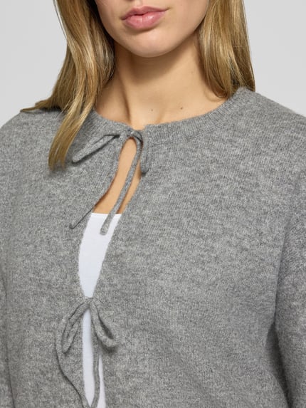 Frauenmodel trägt Strickjacke mit Schleifenverschluss Modell 'PCSALLY' von PIECES in grauer Melange-Strickoptik mit Strukturmuster, Nahaufnahme Oberkörper.