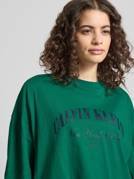 Vrouwenmodel draagt Cropped T-Shirt met Logo-Print van Calvin Klein Jeans in groen, puur katoen, korte fit, ronde hals, logoprint, nahaufnahme bovenlichaam. Ruglengte maat S: 42 cm.