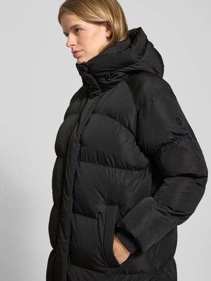 Vrouwenmodel draagt Steppmantel mit Kapuze und Eingrifftaschen van Superdry, zwart, gewatteerd, met kap en insteekzakken, detailopname bovenlichaam. Regular Fit, verborgen ritssluiting, ruglengte 108 cm, schouderbreedte 49 cm, borstwijdte 61 cm, mouwlengte 61 cm.