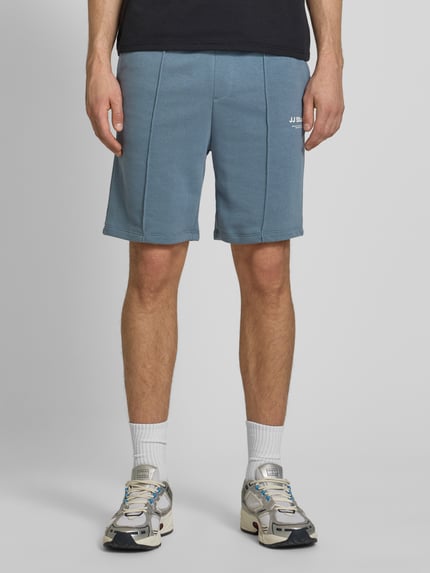 Männermodel trägt Sweatshorts mit Logo Modell 'KANE' von Jack & Jones aus Baumwoll-Mix, Relaxed-Fit, blau, Ganzkörperansicht von vorne.