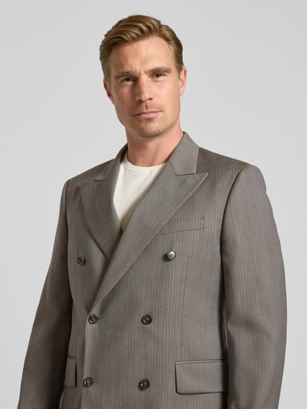 Mannelijk model draagt Sakko aus Wolle mit Label-Patch und Pattentaschen van Tiger of Sweden in lichtgrijs met fijne strepen, regular fit, reverskraag, met dubbele rij knopen, paspelzak op de borst en zijstrookzakken, detailopname bovenlijf. Ruglengte maat 50: 78 cm, schouderbreedte: 50 cm, mouwlengte: 68 cm.