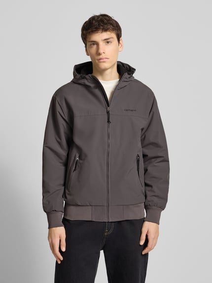 Männermodel trägt Jacke mit Kapuze von Carhartt Work In Progress in Grau, gerippte Abschlüsse, Reißverschlusstaschen, Ganzkörperansicht von vorne.