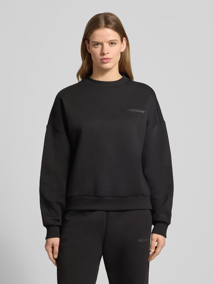 Frauenmodel trägt schwarzen Basic Sweater mit Rundhalsausschnitt und Bündchen am Ärmel von Review, Oversized, Label-Print, Logo-Stickerei, Ganzkörperansicht von vorne.