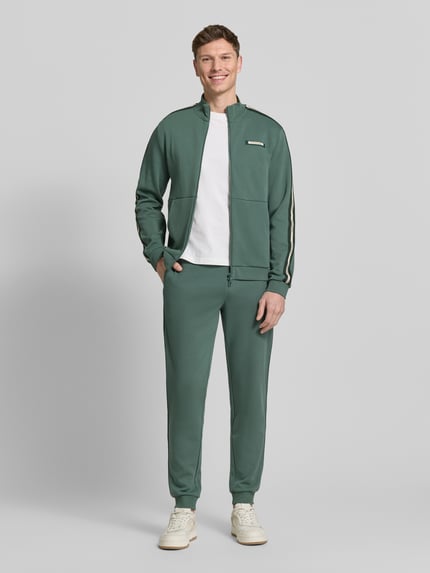 Mannenmodel draagt 'Sweatjacke mit Stehkragen und Zweiwege-Reißverschluss' van EA7 Emporio Armani in regular fit, groengrijs met contrasterende biezen en label-detail, zichtbare zakken en dubbele rits, rechte pijpen, volledige voorkant weergave.