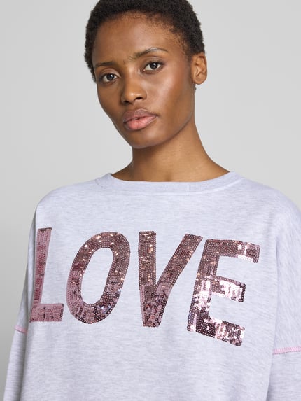 Frauenmodel trägt Oversized Sweatshirt aus reiner Baumwolle Modell 'Love' von miss goodlife in Hellgrau mit Statement-Print und Paillettenbesatz, Nahaufnahme Oberkörper.