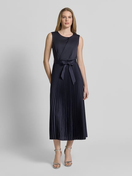 Frauenmodel trägt Cocktailkleid mit Plisseefalten von Vera Mont in Dunkelblau mit Taillenband, Rundhalsausschnitt, Midilänge, Ganzkörperansicht von vorne.