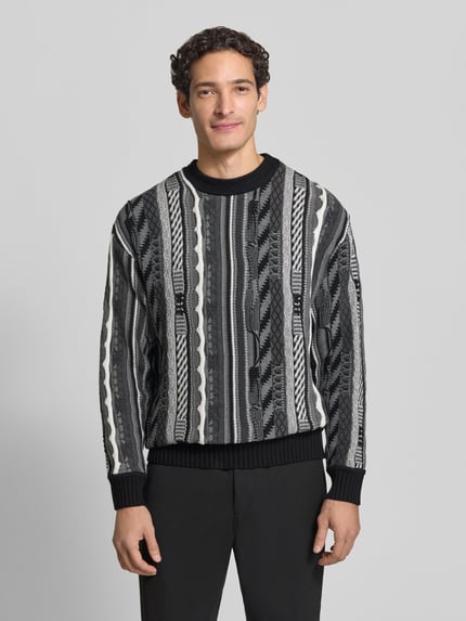 Männermodel trägt Regular Fit Strickpullover aus reiner Baumwolle von COLUCCI, schwarz-grau gemustert, gerippte Abschlüsse, Nahaufnahme Oberkörper.