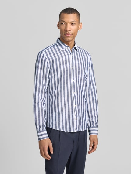 Męski model nosi Slim Fit Business-Hemd mit Kentkragen Modell 'Pit' von JOOP! Collection, biała i niebieska w pionowe pasy, mieszanka lnu z bawełną, krój slim fit, z listwą guzikową i sportowymi mankietami, zaokrąglony dół, długość tyłu w rozmiarze 40: 74 cm, szerokość ramion w rozmiarze 40: 41 cm, długość rękawa w rozmiarze 40: 67 cm, widok całej sylwetki z przodu.