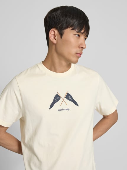 Męski model ma na sobie Regular Fit T-Shirt mit Logo-Print od Forét z czystej bawełny, krótkie rękawy, okrągły dekolt, kremowy kolor, nadruk z przodu, długość tyłu w rozmiarze M: 69 cm, Ganzkörperansicht z przodu.
