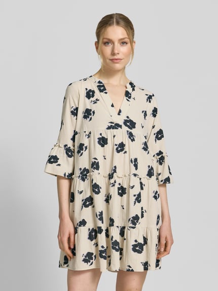 Frauenmodel trägt Ausgestelltes Blusenkleid aus reiner Baumwolle Modell 'DICTHE' von Vero Moda mit Allover-Muster, Henley-Ausschnitt und Trompetenärmeln, cremefarbene Grundfarbe mit dunkelblauen floralen Formen, strukturierte Baumwolltextur, Ganzkörperansicht von vorne.