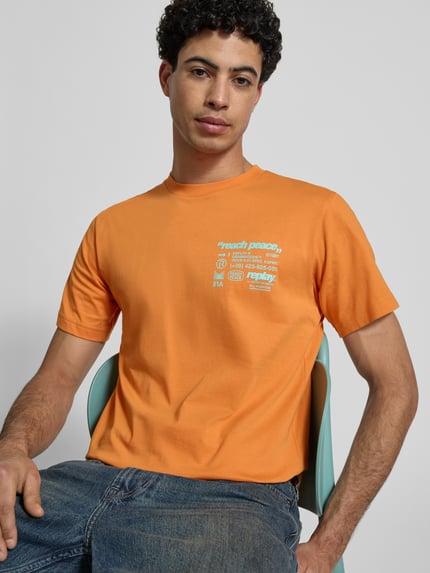 Männermodel trägt Regular Fit T-Shirt mit Statement-Print von Replay in oranje, ronde hals, katoen, korte mouw, tekstprint, nahaufnahme bovenlichaam.