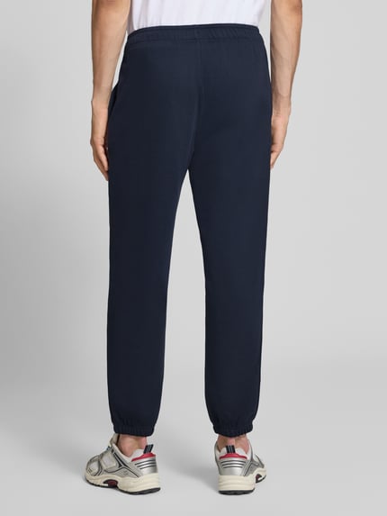 Męski model nosi spodnie dresowe z mieszanki bawełny Champion Sweatpants mit Label-Stitching Modell 'ICONS', prosty krój, elastyczny pas, boczne wpuszczane kieszenie, długość wewnętrzna nogawki w rozmiarze M: 76 cm, ciemnoniebieskie, gładka faktura, widok całej sylwetki od tyłu.