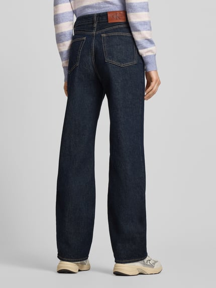 Kobiece model prezentuje Straight Fit Jeansy z detalem z logo od Calvin Klein Jeans, bawełna, model THE STRAIGHT BOOT RESIN, ciemnoniebieskie, szerokość w pasie 37 cm, długość wewnętrzna nogawki 80 cm, widok całej sylwetki z tyłu.