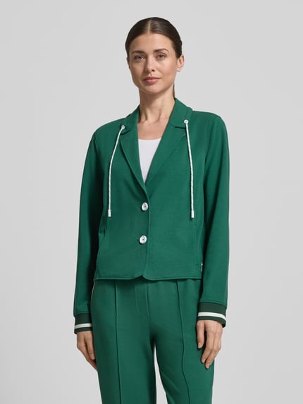 Frauenmodel trägt Regular Fit Blazer mit Eingrifftaschen von Marc Cain in sattem Grün, Baumwoll-Modal-Mix mit Stretch-Anteil, Reverskragen mit Tunnelzug und Knopfverschluss, Ganzkörperansicht von vorne. Rückenlänge bei Größe 36: 55 cm, Brustweite 49 cm, Ärmellänge 63 cm, elastische Ärmelabschlüsse.