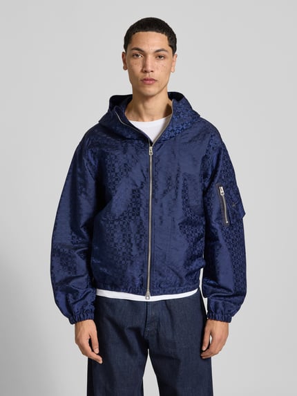 Männermodel trägt Regular Fit Windbreaker mit Zweiwege-Reißverschluss von Kenzo in Dunkelblau, leichte Struktur, elastische Abschlüsse, Ganzkörperansicht von vorne. Herren-Windbreaker, Kapuze, Ärmeltasche, Rückenlänge 68 cm, Schulterbreite 54 cm, Ärmellänge 64 cm.