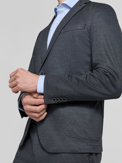 Męski model nosi Regular Fit Anzugsakko mit Viskose-Anteil von s.Oliver BLACK LABEL, wzór allover, krój regular fit, klapy z rewersami, zapięcie na dwa guziki, długi rękaw z kissing buttons, kieszeń na piersi, kieszeń wewnętrzna, naszywane kieszenie, odcień grafitowy, wyraźna faktura, zbliżenie na górną część ciała, zdjęcie z przodu.