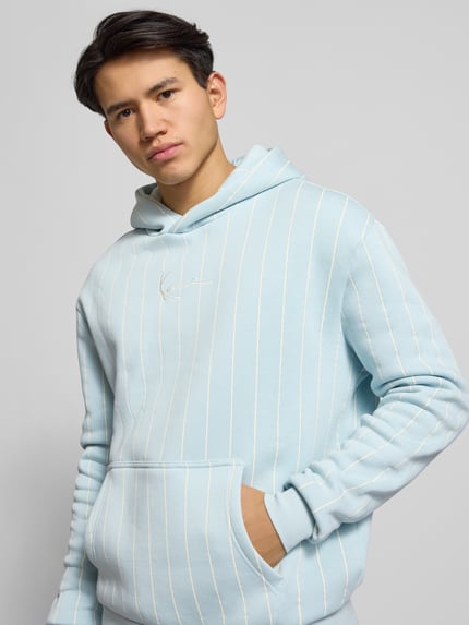 Männermodel draagt Hoodie met kapuze van KARL KANI in lichtblauw met witte strepen, kangoeroezak, geribbelde boorden, katoenmix. Nahaufnahme bovenlichaam.